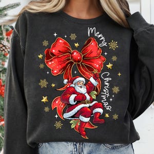 May include: Black sweatshirt featuring a festive design with a large red bow and Santa Claus. The words "Merry Christmas" are written in white script. Gold stars and snowflakes add to the holiday theme.