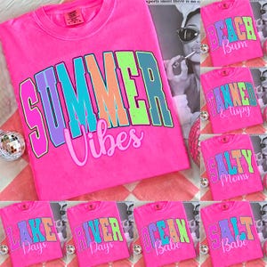 Op de afbeelding: Felroze t-shirts met diverse zomerse slogans in kleurrijke blokletters. Eén shirt toont "SUMMER Vibes", andere tonen zinnen als "BEACH Bum" en "SALTY Moms". De shirts liggen op een rood-wit geruit oppervlak.
