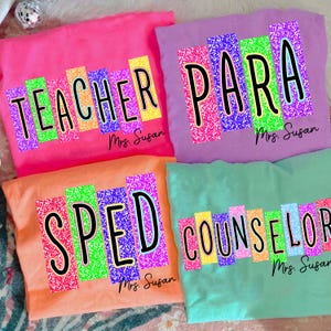 May include: Four colorful t-shirts with text. The shirts are pink, purple, orange, and teal. Each shirt has a different title in large, colorful letters: TEACHER, PARA, SPED, and COUNSELOR. The name "Mrs. Susan" is printed below each title.