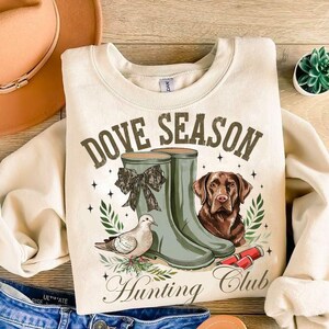 May include: A beige sweatshirt with the text "Dove Season Hunting Club" in brown lettering. The design features a pair of green rubber boots, a brown dog, and a dove.