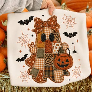 May include: A white sweatshirt featuring a patchwork ghost graphic with a large bow. The ghost is made of various brown, orange, and green fabric patterns, holding a jack-o'-lantern bucket. The design includes black bats, spiderwebs, and stars.