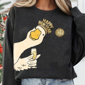 May include: Black sweatshirt featuring a cartoon goose wearing a "Happy New Year" crown and holding a champagne glass. A gold disco ball hangs nearby. The design is in shades of gold, white, and black.