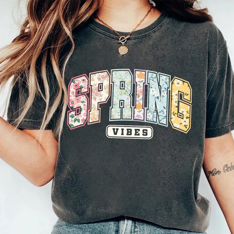 Puede incluir: Camiseta gris oscuro con las palabras "SPRING VIBES". La palabra "SPRING" tiene un dise&ntilde;o floral en un estilo universitario. "VIBES" est&aacute; en un rect&aacute;ngulo blanco. Una prenda casual para el d&iacute;a a d&iacute;a.