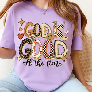 May include: A lilac-colored t-shirt with the phrase "GOD IS GOOD all the time" in a mix of fonts and patterns. The design includes leopard print, checkerboard, and floral elements, along with a smiley face and heart shapes.