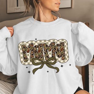May include: White sweatshirt featuring the word "MAMA" in a camouflage design, set against a checkered background. A green bow sits beneath the word, with small black star accents. The sweatshirt is a relaxed fit.