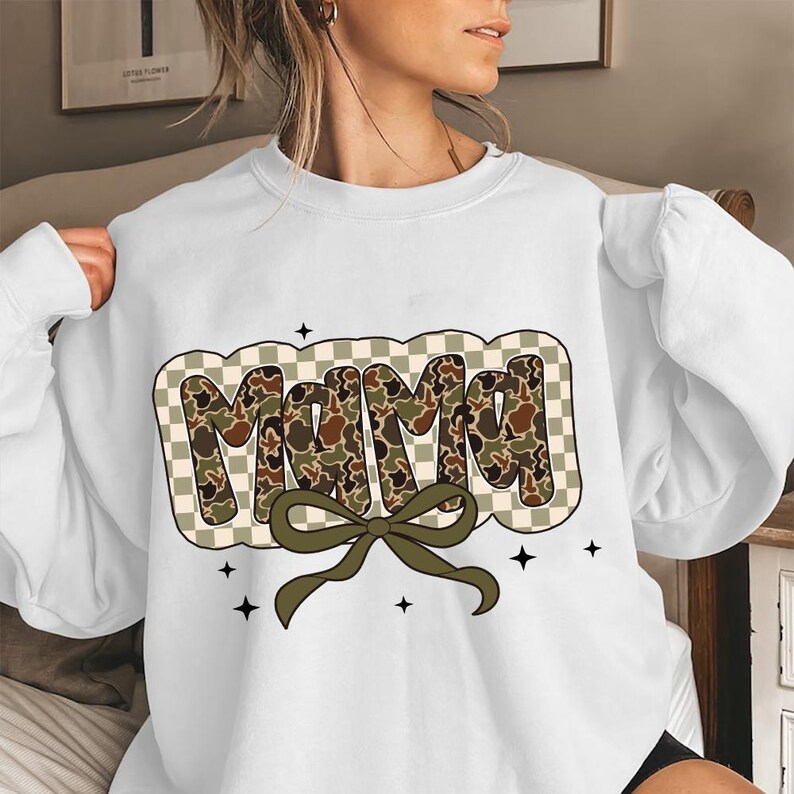 May include: White sweatshirt featuring the word "MAMA" in a camouflage design, set against a checkered background. A green bow sits beneath the word, with small black star accents. The sweatshirt is a relaxed fit.