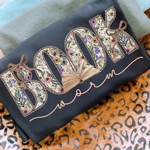 K&ouml;nnte beinhalten: Dunkelgraues T-Shirt mit den Worten "BOOK worm" in einem floralen Design. Die Buchstaben sind mit bunten Blumen gef&uuml;llt und das Wort "worm" ist in einer Schreibschrift geschrieben. Ein braunes offenes Buch befindet sich in der Mitte.