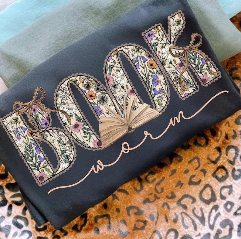 K&ouml;nnte beinhalten: Dunkelgraues T-Shirt mit den Worten "BOOK worm" in einem floralen Design. Die Buchstaben sind mit bunten Blumen gef&uuml;llt und das Wort "worm" ist in einer Schreibschrift geschrieben. Ein braunes offenes Buch befindet sich in der Mitte.