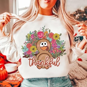 May include: White sweatshirt with a brown turkey surrounded by colorful flowers. The turkey has a pink beak and a pink tongue sticking out. The text "thankful" is written below the turkey.
