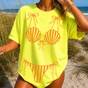 May include: A bright yellow t-shirt with an orange seashell bikini design. The bikini top has two seashell cups with vertical lines and bows. The bikini bottom has vertical stripes and bows. The shirt is casual beachwear.