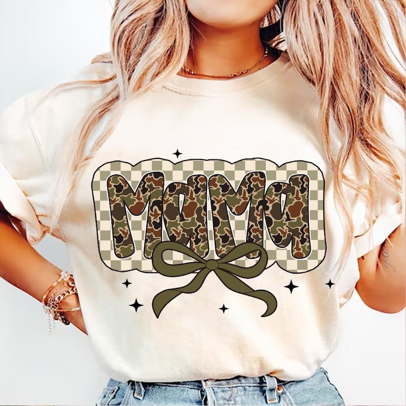 May include: A cream-coloured sweatshirt featuring the word "Mama" in a camouflage pattern, framed by a checkered design. A green bow sits below the word, with four black star accents.