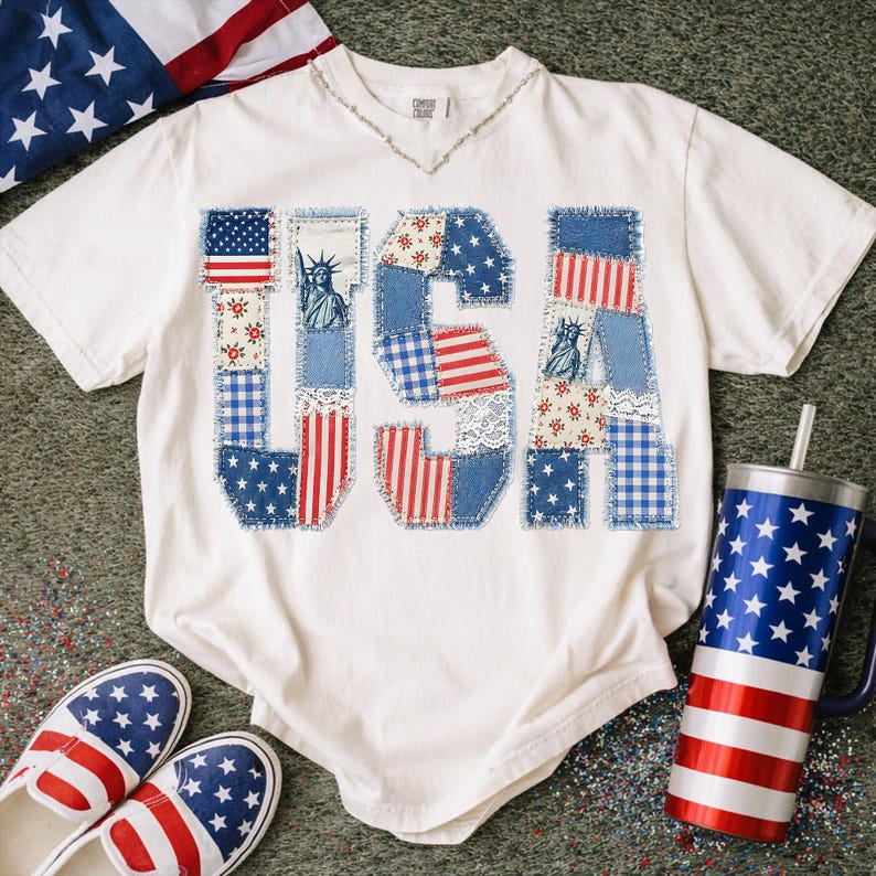 K&ouml;nnte beinhalten: Wei&szlig;es T-Shirt mit dem Wort "USA" im Patchwork-Stil, mit Designs der amerikanischen Flagge und der Freiheitsstatue. Enth&auml;lt passende Schuhe und einen Becher mit patriotischem Thema. Eine amerikanische Flagge ist im Hintergrund.