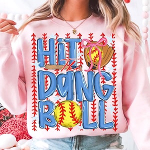 Può includere: Felpa rosa chiaro con la scritta "Hit the Dang Ball" in lettere blu a bolle. Il design include una palla da baseball, un guanto e elementi decorativi. Uno stile casual e confortevole.