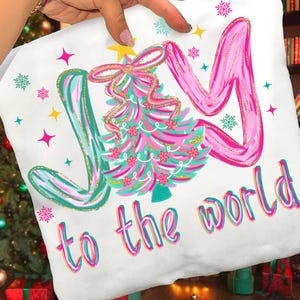 May include: White Christmas-themed pillow with the words "JOY to the world" in a colorful, whimsical font. The design includes a pink and teal Christmas tree with a gold star and bow, surrounded by stars and snowflakes.