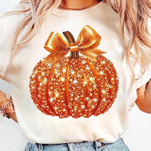 Può includere: T-shirt color crema con un design di zucca arancione scintillante. La zucca è adornata con glitter e un grande fiocco. Il design è centrato sulla maglietta, creando un look festoso e accattivante.