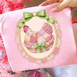 May include: A pink pillow with a decorative hen design. The hen is made of patchwork fabric in shades of pink, floral patterns, and hearts. A green bow adorns the top and bottom of the oval frame. The pillow is held by a person.