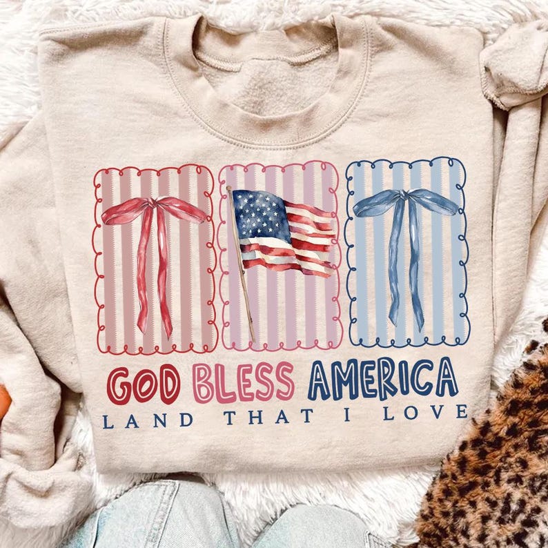 May include: Beige sweatshirt with a patriotic design. Features three framed images: a red bow, an American flag, and a blue bow. The text "GOD BLESS AMERICA" and "LAND THAT I LOVE" are printed in red and blue.