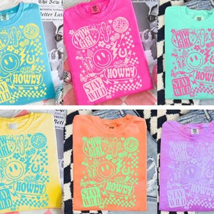 May include: Six colorful t-shirts with the text "Cow Girl", "Howdy", and "Stay Wild" in a playful design. The shirts are in shades of blue, pink, green, yellow, orange, and purple. The design includes a smiley face, cactus, and disco ball.