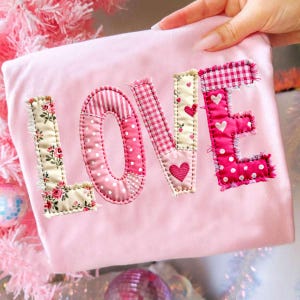Pode incluir: Um tecido rosa claro com a palavra "LOVE" aplicada em vários padrões rosa. As letras são costuradas com tecidos florais, de bolinhas, listrados e xadrez, com detalhes de coração. As bordas das letras têm uma aparência desfiada e texturizada.