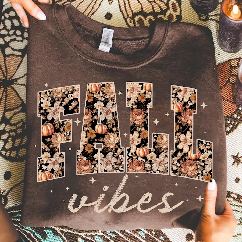 May include: A brown sweatshirt with the words "FALL vibes" in a floral design. The letters are filled with pumpkins and flowers in shades of orange, cream, and brown. The sweatshirt is held up by two hands.