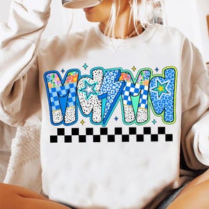 Peut inclure: Sweat-shirt blanc avec le mot "MAMA" dans un design rétro coloré. Les lettres présentent des motifs floraux, léopard et à carreaux en bleu, vert et blanc, avec une bordure à carreaux noir et blanc.