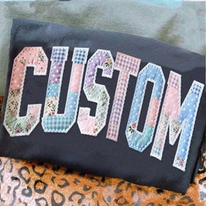 May include: A dark gray t-shirt with the word "CUSTOM" appliqu&eacute;d across the front. The letters are made of patchwork fabric in various pastel colors and patterns, including floral, polka dot, and gingham. The letters are outlined in white.