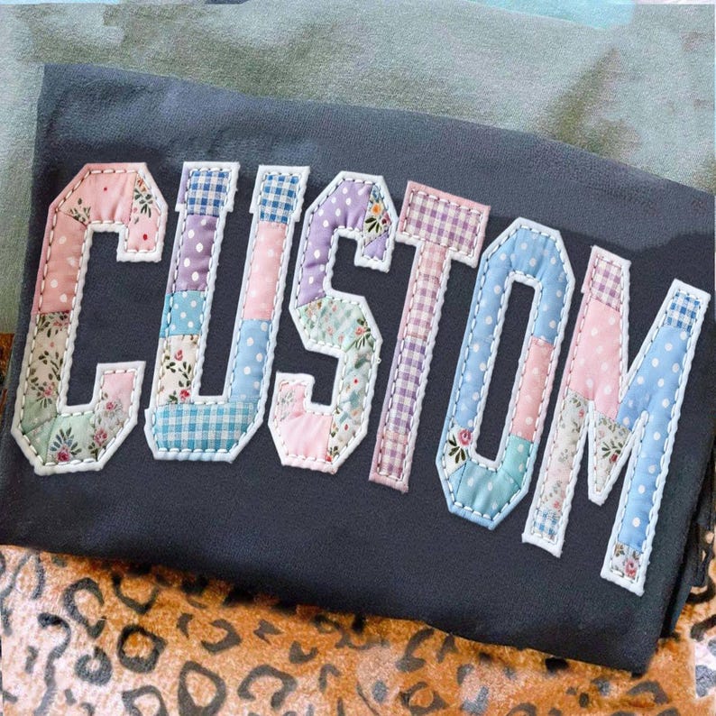May include: A dark gray t-shirt with the word "CUSTOM" appliqu&eacute;d across the front. The letters are made of patchwork fabric in various pastel colors and patterns, including floral, polka dot, and gingham. The letters are outlined in white.