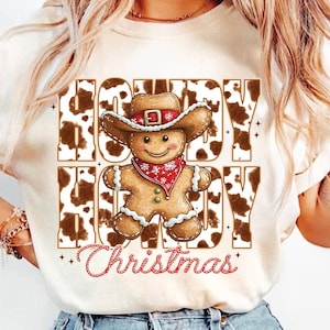 May include: White t-shirt with a brown and white cow print design. The shirt features a gingerbread man wearing a cowboy hat and the text "Howdy Hoody Christmas".