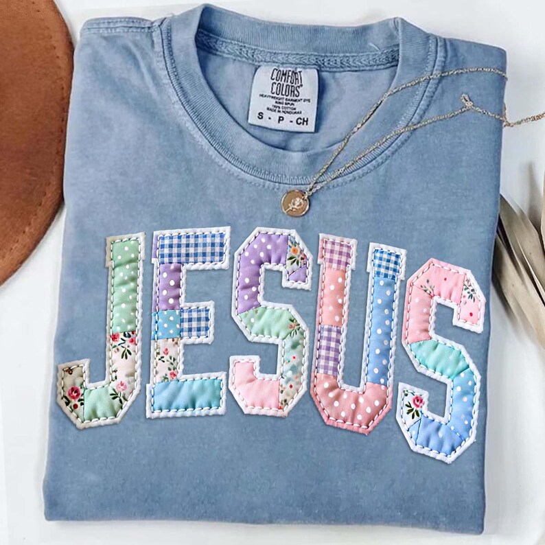May include: A light blue Comfort Colors t-shirt with the word "JESUS" appliqued across the front. The letters are made of patchwork fabric in various pastel colors and patterns. A gold necklace with a pendant is draped over the shirt.