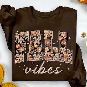 May include: A brown sweatshirt with the words "FALL vibes" in a floral design. The letters are filled with a pattern of flowers and pumpkins. The word "vibes" is written in a cursive font.