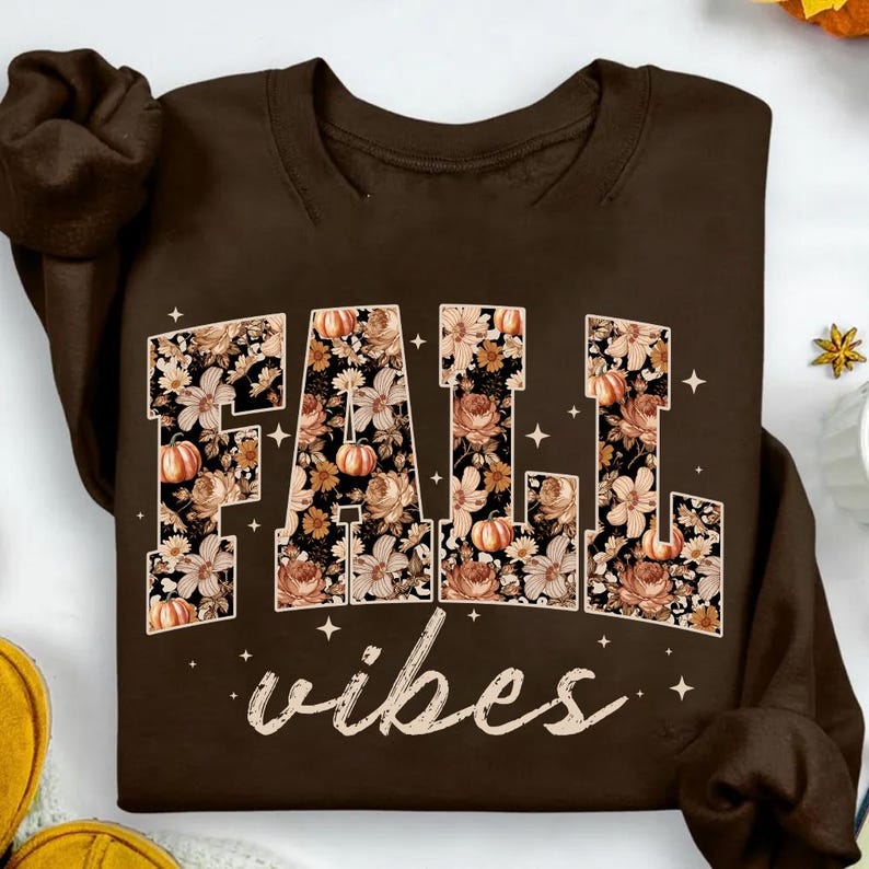 May include: A brown sweatshirt with the words "FALL vibes" in a floral design. The letters are filled with a pattern of flowers and pumpkins. The word "vibes" is written in a cursive font.