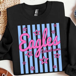 Puede incluir: Sudadera negra con un gr&aacute;fico rosa y azul. El gr&aacute;fico presenta la palabra "Eagles" en escritura rosa, rayas verticales azules y formas de estrella rosa. Tambi&eacute;n son visibles las palabras "School name".