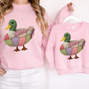 May include: Pink sweatshirts featuring a patchwork duck design. The duck has a green head, yellow beak, and a body made of various fabric patterns. The sweatshirts are a matching set, one adult and one child size.