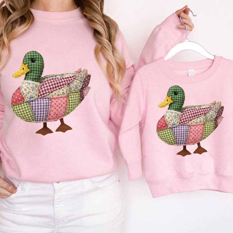 May include: Pink sweatshirts featuring a patchwork duck design. The duck has a green head, yellow beak, and a body made of various fabric patterns. The sweatshirts are a matching set, one adult and one child size.