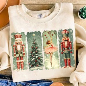 May include: A cream-coloured sweatshirt with a vintage-style Christmas design. The design features three panels with a nutcracker, a Christmas tree, and a ballerina. The panels are surrounded by a distressed white border.