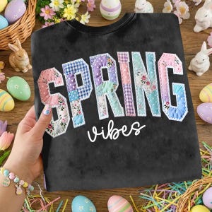 May include: Black sweatshirt with the words "SPRING vibes" in a patchwork design. The letters are in various pastel colors and floral patterns. The image is surrounded by Easter decorations, including eggs and bunnies.