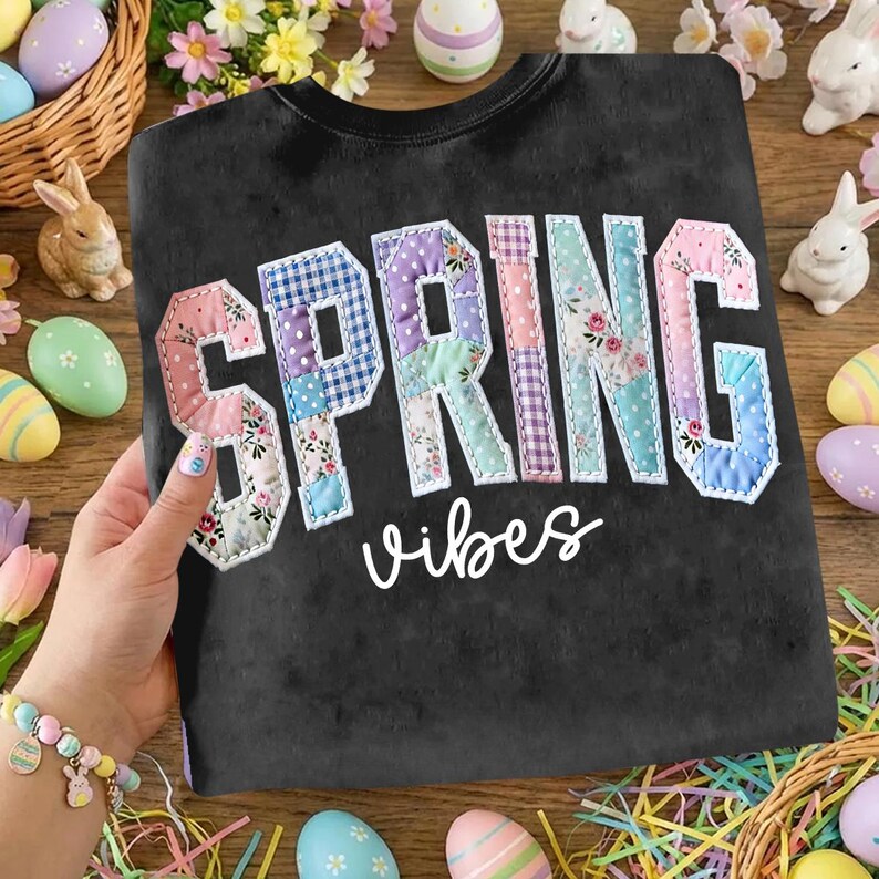 May include: Black sweatshirt with the words "SPRING vibes" in a patchwork design. The letters are in various pastel colors and floral patterns. The image is surrounded by Easter decorations, including eggs and bunnies.