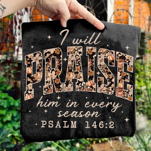 May include: Black t-shirt with the text "I will PRAISE him in every season" and "PSALM 146:2". The word "PRAISE" is filled with a floral and pumpkin design. The shirt is held up by a hand.