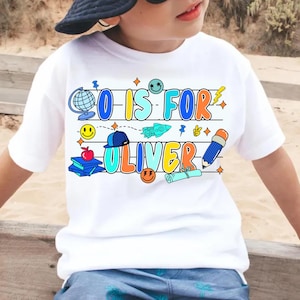 May include: White t-shirt with the text "O IS FOR OLIVER" in colorful, playful lettering. The design includes a globe, smiley faces, a rocket ship, a pencil, and other school-themed graphics. The shirt is worn by a child.