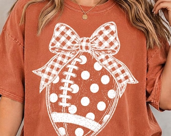 May include: A rust-colored short-sleeve t-shirt featuring a large white graphic print of an American football decorated with polka dots and topped with a prominent gingham-patterned bow. The design is centered on the front of the garment.