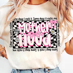 Peut inclure: T-shirt cr&egrave;me avec un motif rose et blanc "Motherhood" sur fond damier noir et blanc. Le motif comprend des n&oelig;uds roses, des fleurs et les phrases "Either way we're rockin'" et "Some days I rock it. Some days it rocks me."