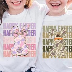 May include: Two white t-shirts with a graphic of a bunny rabbit. The bunny on the left is pink and purple with a bow and the text "Egg Hunter". The bunny on the right is camouflage with a hat and the text "Egg Hunter". Both shirts have the text "Happy Easter" repeated multiple times.