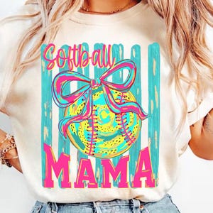 May include: Cream-colored t-shirt featuring a softball design. The design includes a softball with a pink and blue bow, set against a backdrop of teal stripes. The word "Softball" is written above the ball, and "MAMA" below.