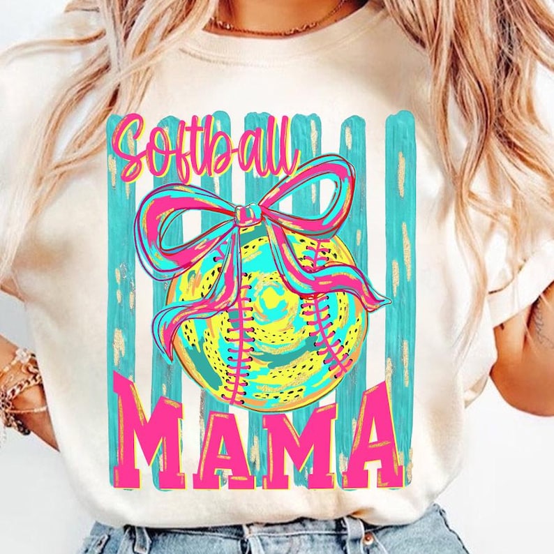May include: Cream-colored t-shirt featuring a softball design. The design includes a softball with a pink and blue bow, set against a backdrop of teal stripes. The word "Softball" is written above the ball, and "MAMA" below.