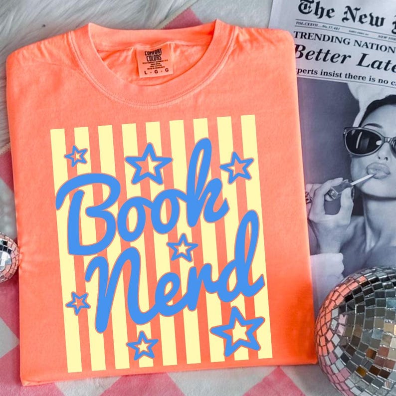 May include: Coral-colored t-shirt with the words "Book Nerd" in blue script, surrounded by blue stars. The design is set against yellow vertical stripes. The shirt is folded and displayed on a pink and white checkered surface.