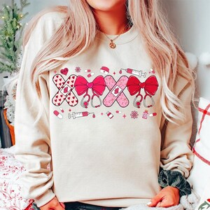 May include: A cream-colored sweatshirt with a pink and red design featuring band-aids, hearts, bows, stethoscopes, and syringes.