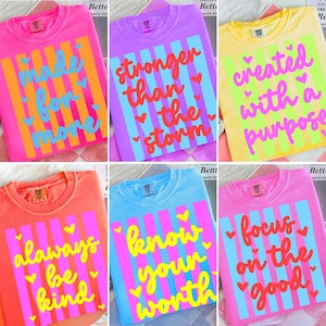 May include: A collection of colorful t-shirts with inspirational quotes. Each shirt features a different message in a vibrant font, set against a striped background. The shirts are in various colors, including pink, purple, and yellow.