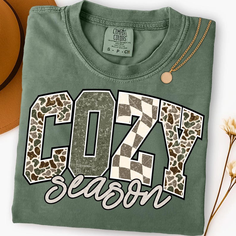 K&ouml;nnte beinhalten: Salbeigr&uuml;nes T-Shirt mit dem Schriftzug "COZY Season" in einem verwitterten, mehrschichtigen Design. Die Buchstaben "COZY" weisen ein Camouflage-Muster und ein Schachbrettmuster auf. Eine goldene Halskette mit rundem Anh&auml;nger liegt auf dem Shirt.
