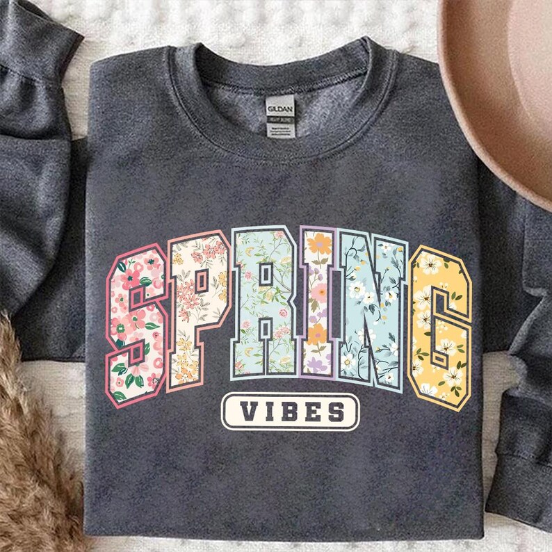 Puede incluir: Una sudadera gris oscuro con las palabras "SPRING VIBES" en un dise&ntilde;o floral. Las letras est&aacute;n rellenas de patrones florales en rosa, azul y amarillo. La sudadera tiene cuello redondo y mangas largas.