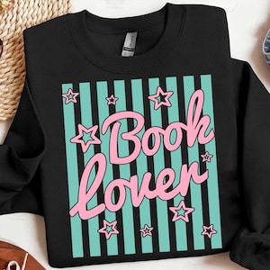 May include: Black sweatshirt with a teal and black striped design. The design features the words "Book Lover" in pink script, surrounded by pink stars. The sweatshirt is made of a soft material.